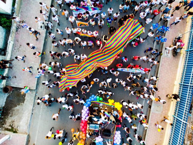 Birds-eye view of a large LGBTQ+ flag in the middle of a pride parade
